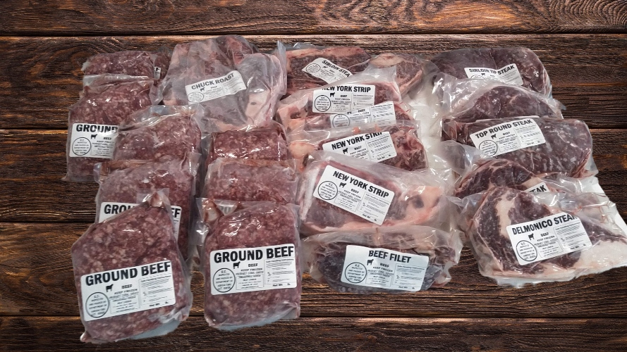 Assortment of packaged beef cuts including ground beef, chuck roast, New York strip, top round steak, beef filet, and Delmonico steak displayed on a wooden surface.