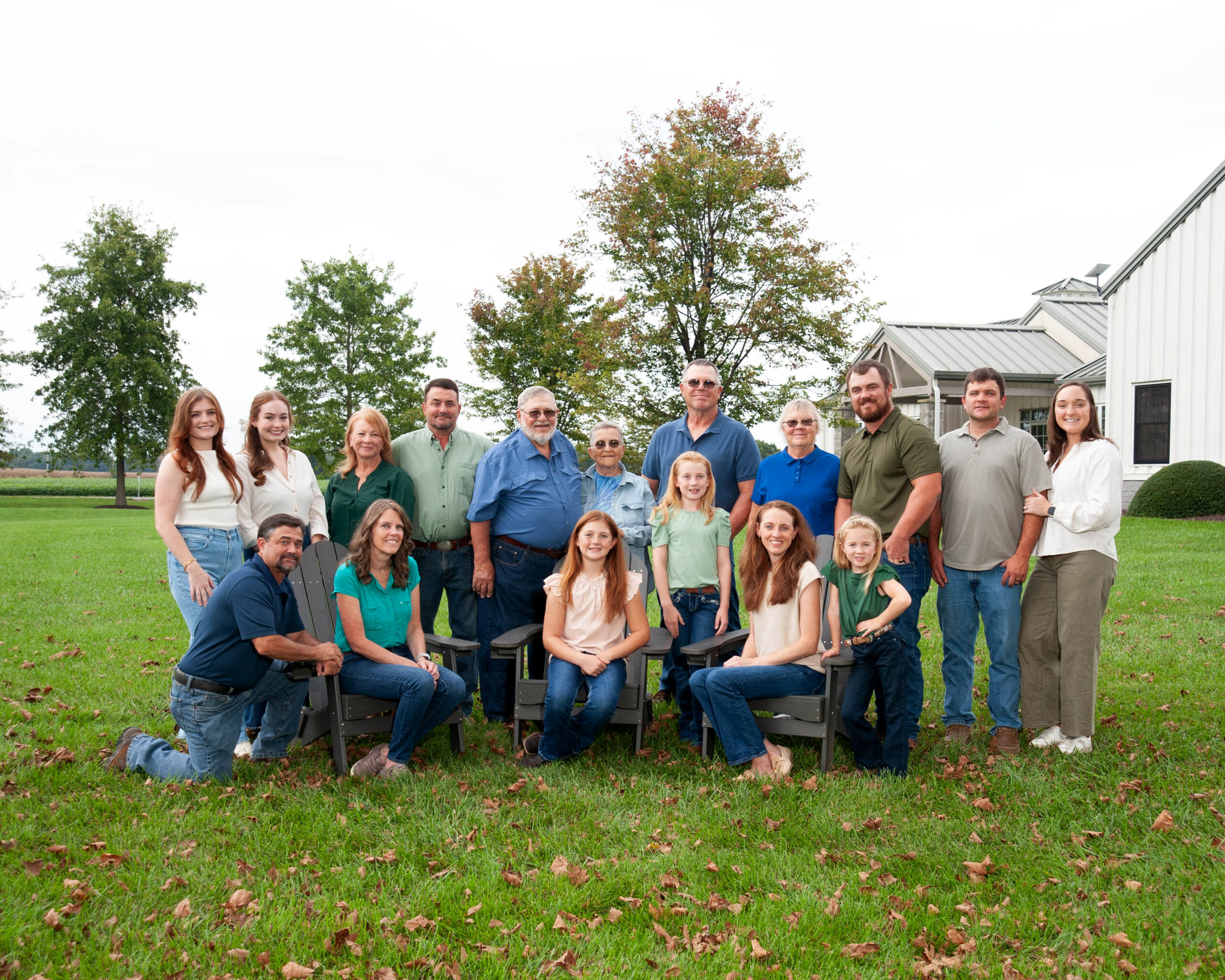 Family reunion photo featuring a diverse group of 20 people standing and sitting in a grassy outdoor setting, with trees and a building in the background. The group includes adults and children, all dressed in casual attire, smiling and posing together.