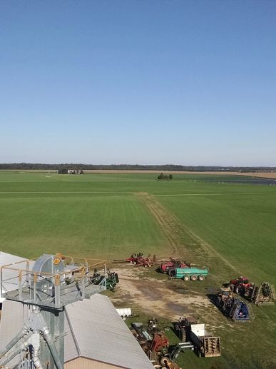 Aerial view of a lush green agricultural field with farm machinery parked nearby, under a clear blue sky. The image captures the expansive landscape, showcasing the farming equipment and the surrounding fields.