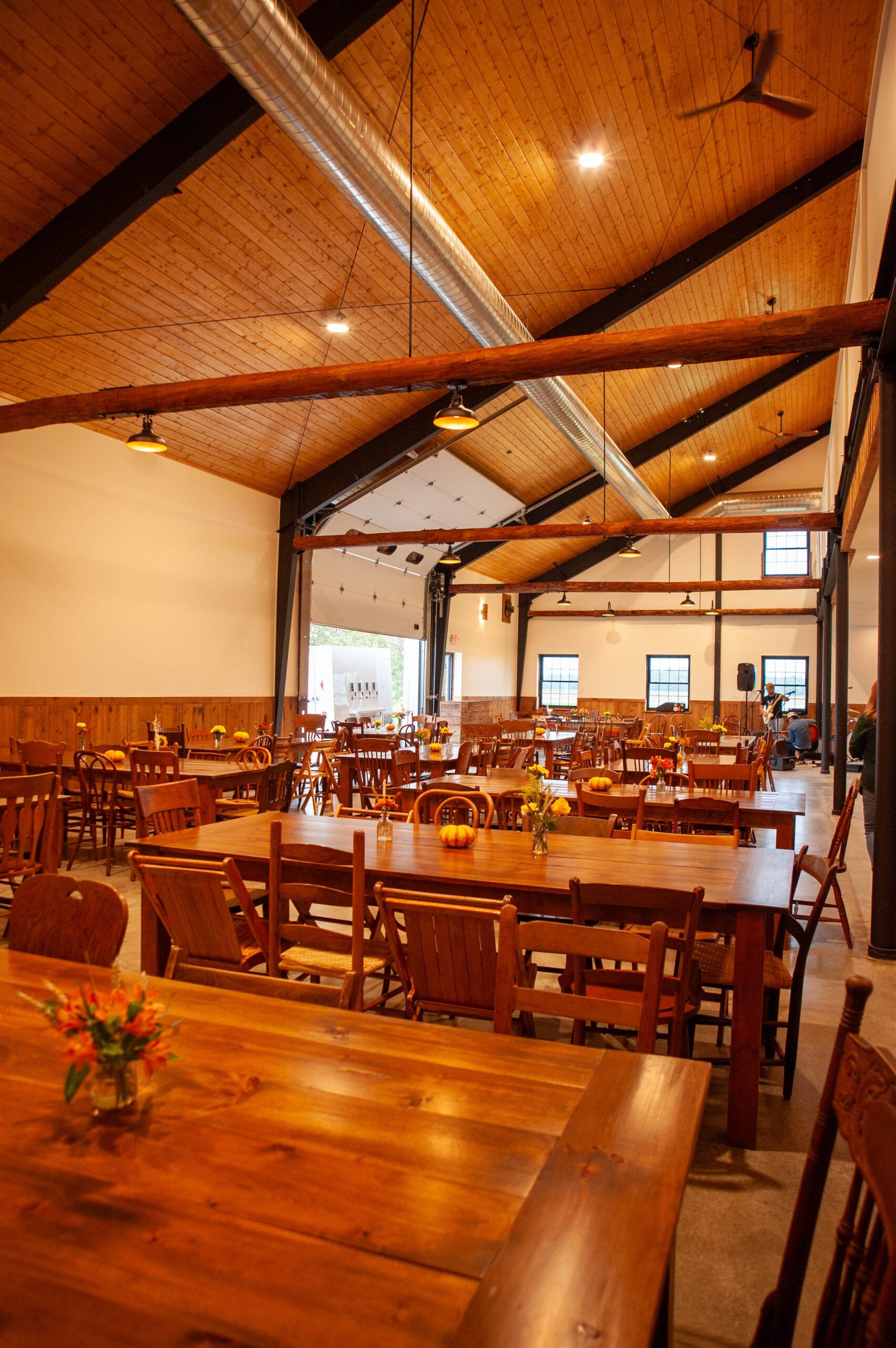 Spacious event venue interior featuring high wooden ceilings, rustic wooden tables, and decorative flower arrangements. The setting is well-lit with hanging lights and offers a warm, inviting atmosphere for gatherings and celebrations.